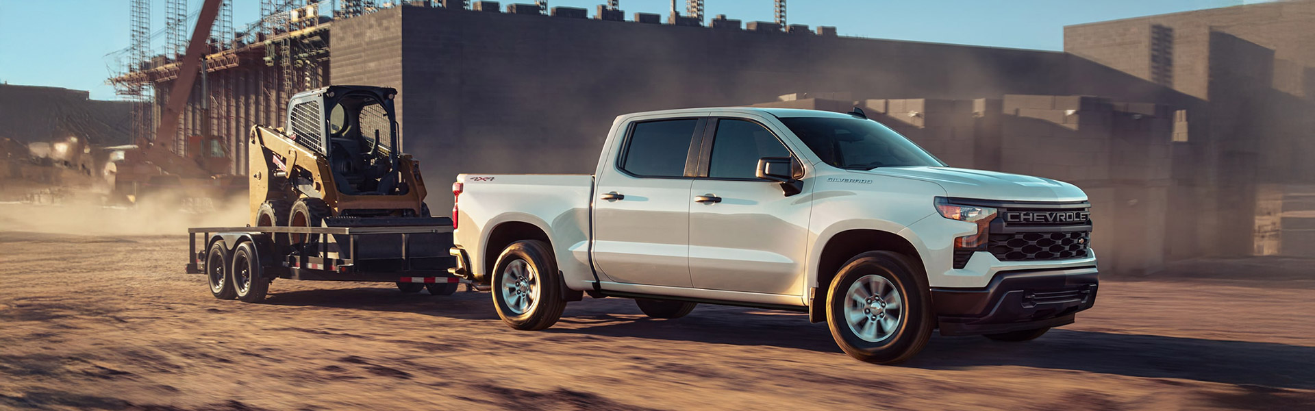 2025 Chevrolet Silverado 1500 pickup truck towing construction equipment on a flatbed trailer at a dusty jobsite.
