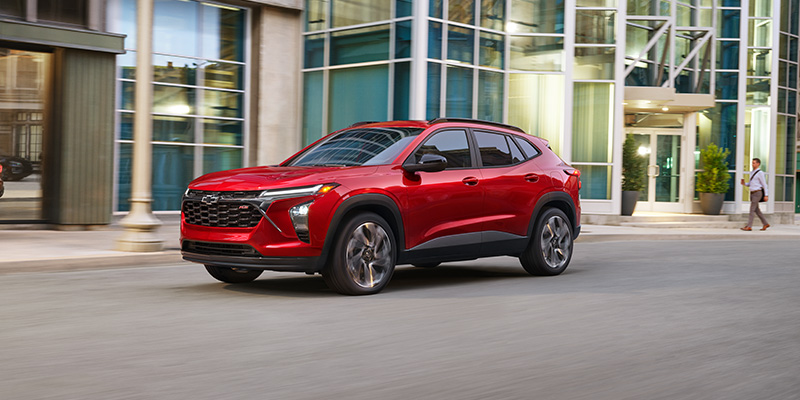 A sleek red Trax drives past modern glass buildings on a city street. The vehicle's design is dynamic, suggesting speed and modernity.