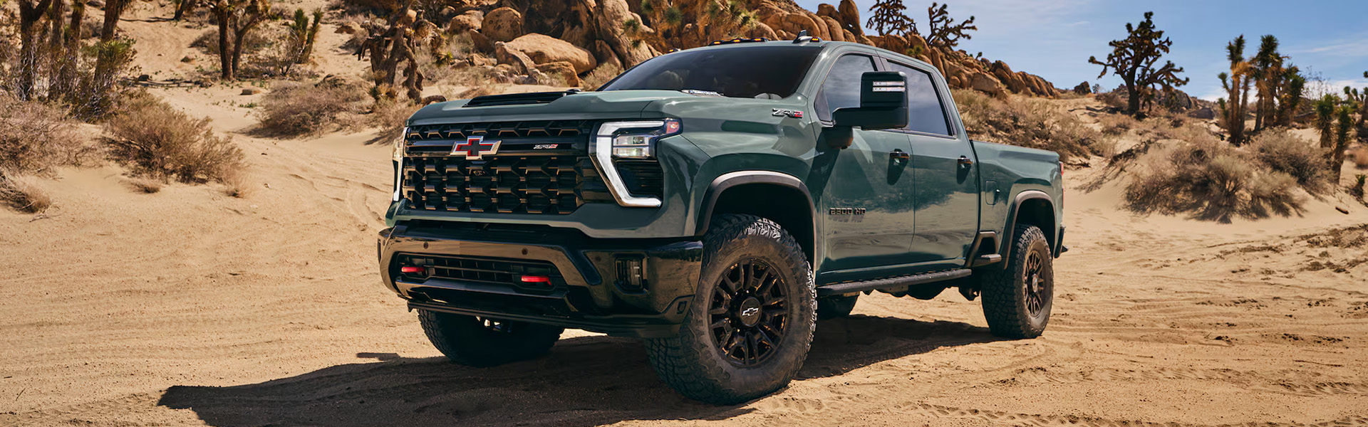 2026 Chevrolet Silverado HD ZR2 heavy-duty pickup truck in green parked on a rugged desert trail, showcasing its off-road stance, aggressive front grille, lifted suspension, and all-terrain tires.